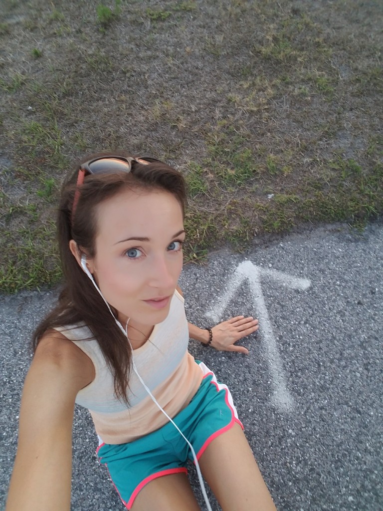 Woman wearing headphones and athletic clothing, sitting on pavement next to a white painted arrow
