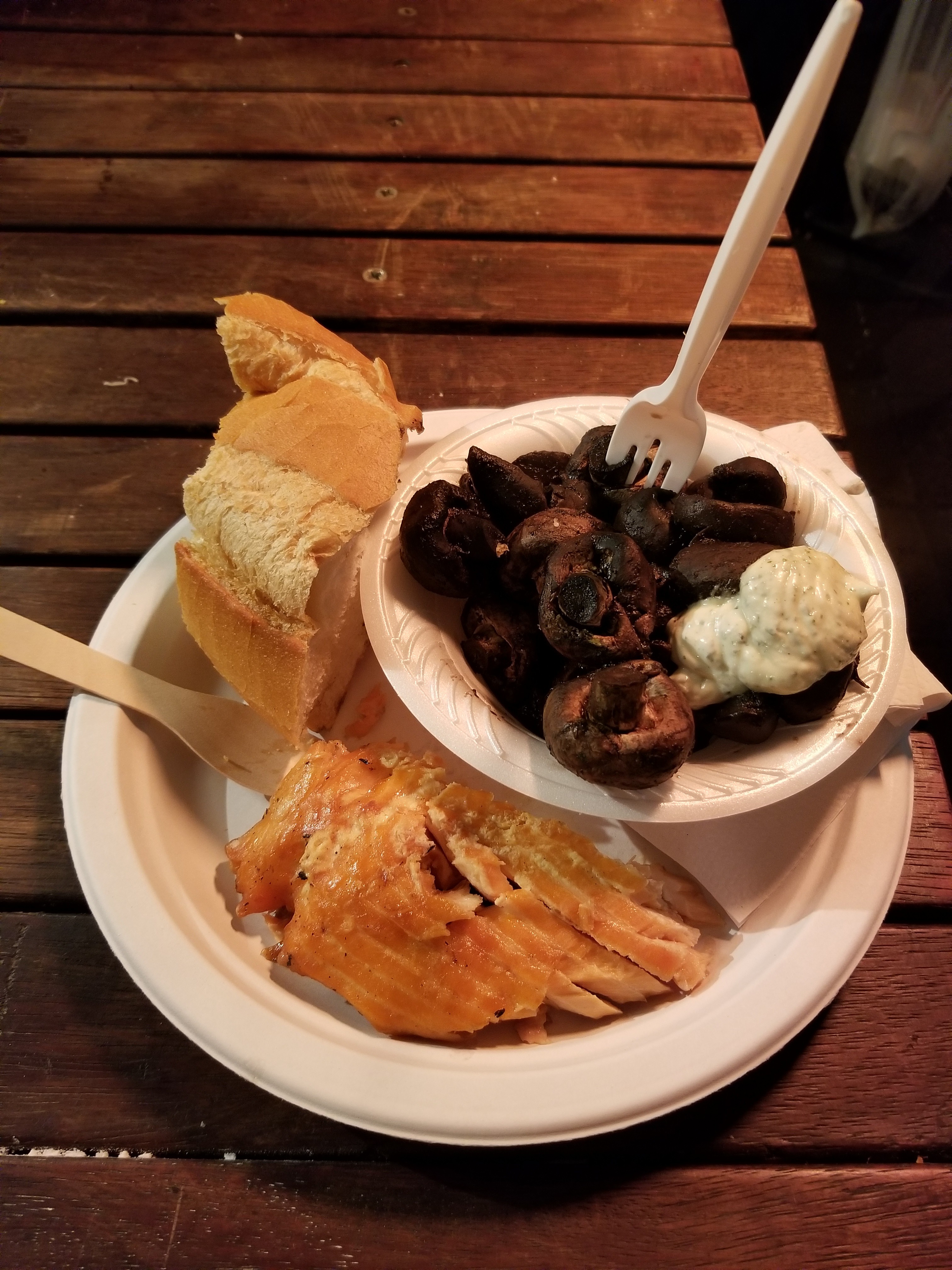 Styrofoam bowl of mushrooms on a paper plate with bread , salmon, and plastic cutlery